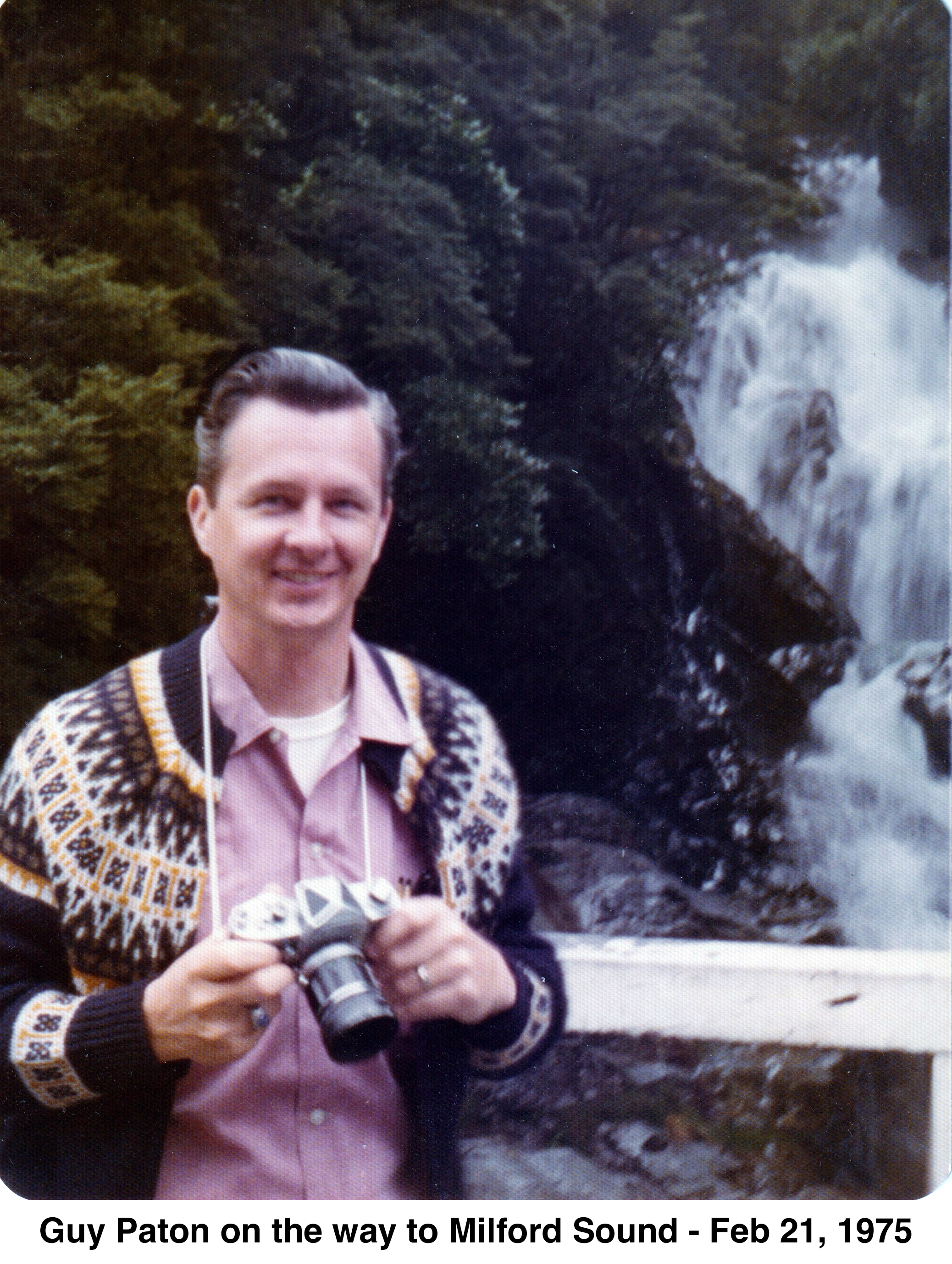 Guy is wearing a sweater and holding a camera in front of green               bushes and a waterfall