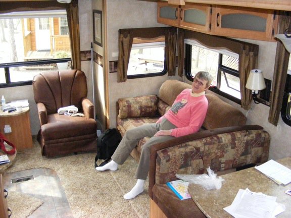 Gail is sitting          on our little couch on the side of the living          area. One of the easy chairs is visible at the          back of the room.