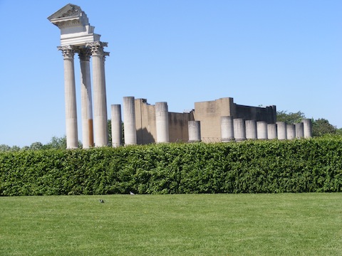 Majestic columns, mostly incomplete, supporting             part of a portico, rise behind a green hedge