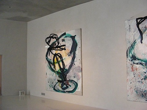 A wall-sized painting consists of swirls of black            and green paint. In the center of the large swirl is            an area of yellow and orange. Outside the swirls are            splotches of grey with, perhaps, a tint of lavender.