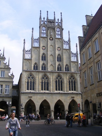 The facade of this building consists of four large open pointed arches           at street level. Above them are four windows in pointed arches of about the            same size. Above them is a front that gets narrower in four square steps to           the top. In this false front are six windows in pointed arches as well           as decorative arches with solid interiors. At the top are two statues.           The whole is made of tan stones.