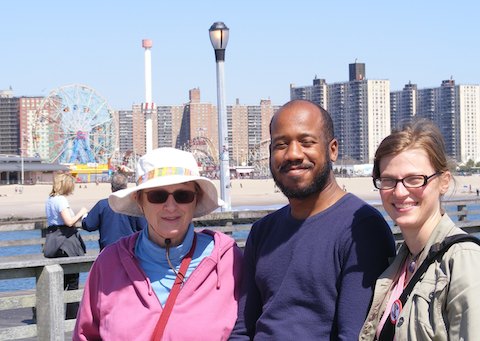 A close up photo of Gail, Eric and CJ with the              ferris wheel and other rides visible beyond the             water and sand. Behind the amusement park are high-rise             buildings.