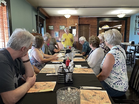 Eunice is standing at the far end of the table with the other             attendees sitting on bothh sides of the table and looking at her