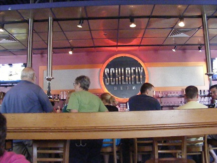 Bob and Gail stand near            the bar. A large neon-lit Schlafly sign hangs on the wall           behind the bar