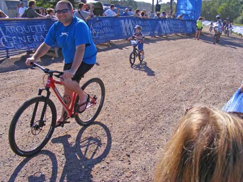 Chris smiles at the camera as he rides
               past the finish line. Little Griffen follows
               him. 